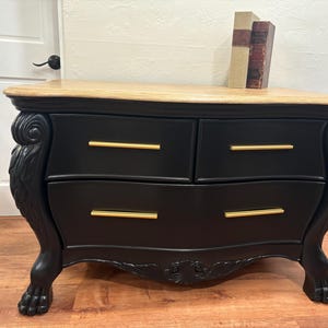 May include: A black dresser with gold handles and ornate lion's paw feet. The dresser has three drawers and a wood top.