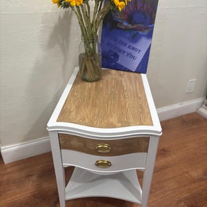 May include: White end table with two drawers and a lower shelf. The table top is wood with a light brown stain. The drawers have gold knobs. A bouquet of sunflowers in a glass jar is on the table, along with a framed print that says "Tie the knot without you".