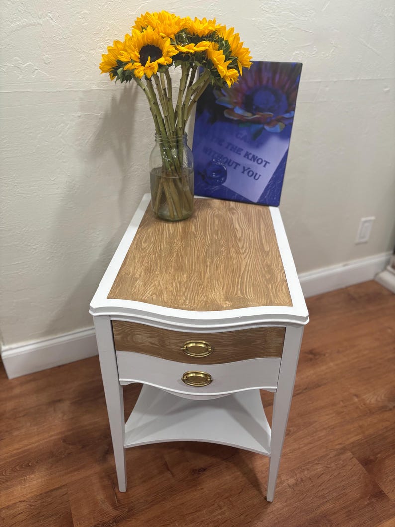 May include: White end table with two drawers and a lower shelf. The table top is wood with a light brown stain. The drawers have gold knobs. A bouquet of sunflowers in a glass jar is on the table, along with a framed print that says "Tie the knot without you".