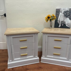 May include: Two white nightstands with three drawers each. The drawers have gold handles. The nightstands have a light wood top and a white base.