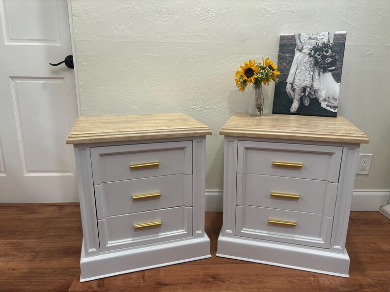 May include: Two white nightstands with three drawers each. The drawers have gold handles. The nightstands have a light wood top and a white base.