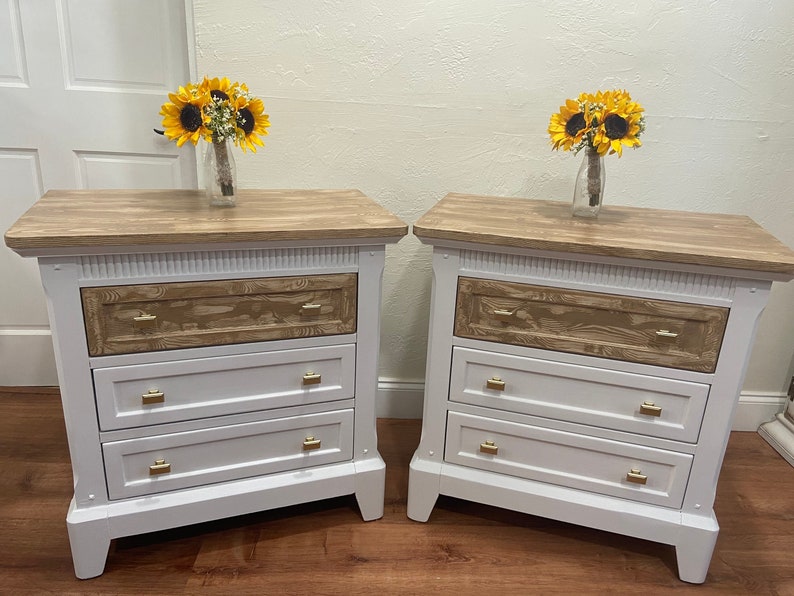 May include: Two white nightstands with three drawers each. The top drawer is wood-toned, and the bottom two drawers are white. The nightstands have gold-colored hardware. Each nightstand is topped with a bouquet of sunflowers.