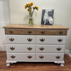 May include: White dresser with eight drawers and gold-toned hardware. The top drawer is made of wood with a different grain than the rest of the dresser. The dresser is sitting on a wood floor.
