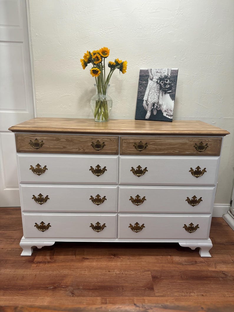 May include: White dresser with eight drawers and gold-toned hardware. The top drawer is made of wood with a different grain than the rest of the dresser. The dresser is sitting on a wood floor.
