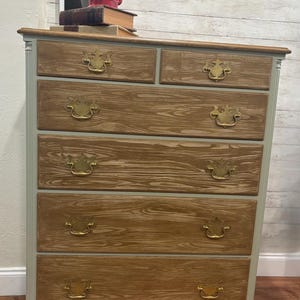 May include: A six-drawer dresser with a light green painted finish and wood grain showing through. The dresser has brass handles with a decorative design. The dresser is sitting on a wooden floor.