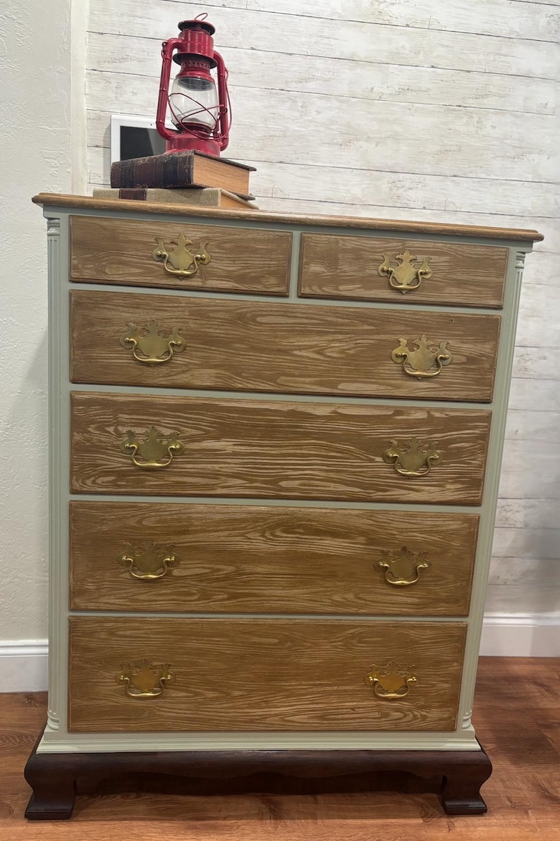 May include: A six-drawer dresser with a light green painted finish and wood grain showing through. The dresser has brass handles with a decorative design. The dresser is sitting on a wooden floor.