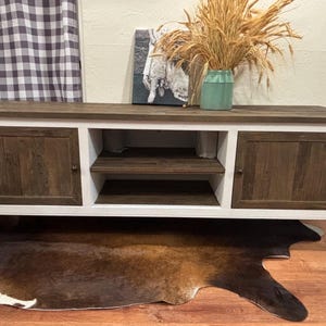 May include: A white and brown wooden TV stand with two doors on each side and a shelf in the middle. The stand has a white base and decorative legs.