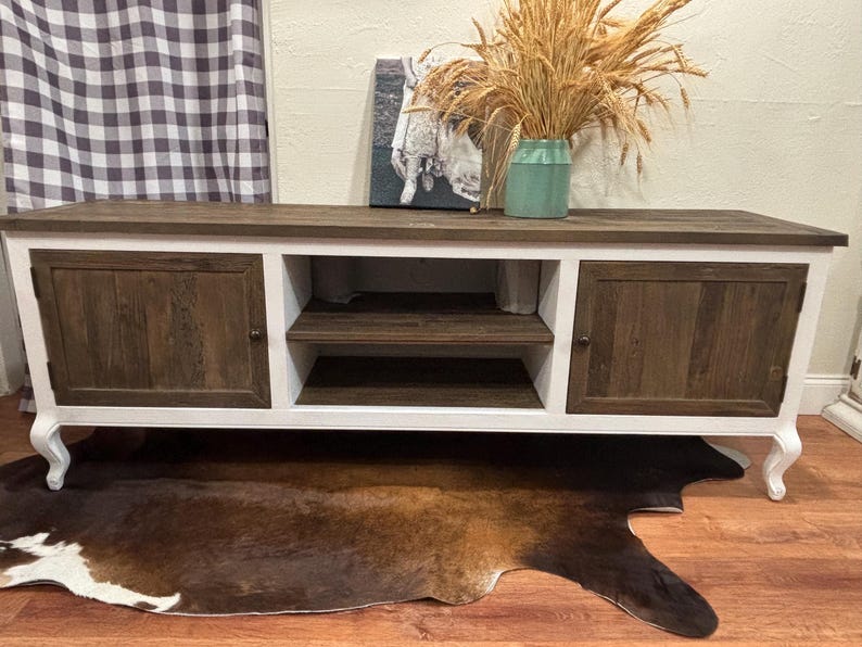 May include: A white and brown wooden TV stand with two doors on each side and a shelf in the middle. The stand has a white base and decorative legs.