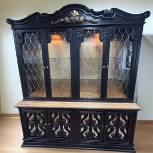 May include: A black wooden hutch with ornate details and gold accents. The hutch has glass doors with a wire mesh pattern and a lower section with three doors. The doors have a decorative cutout design.