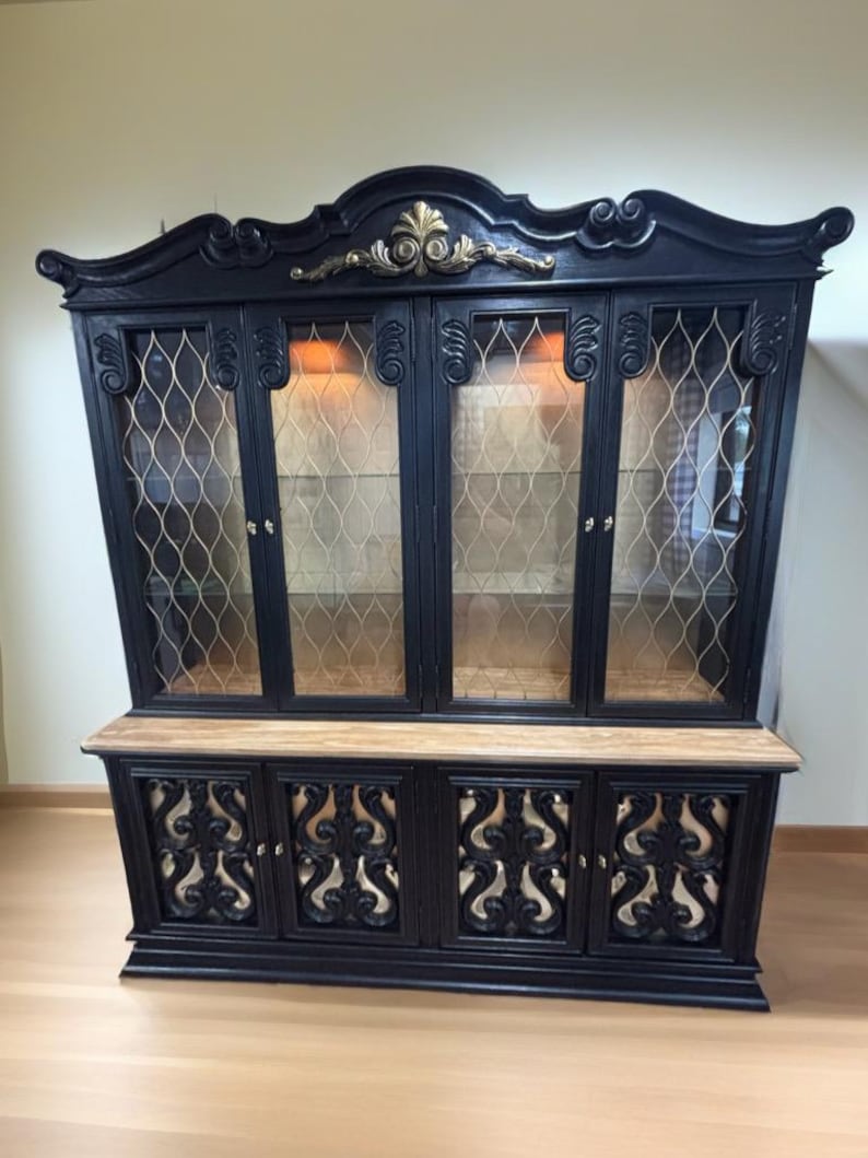 May include: A black wooden hutch with ornate details and gold accents. The hutch has glass doors with a wire mesh pattern and a lower section with three doors. The doors have a decorative cutout design.