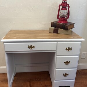 May include: White desk with four drawers and a wood top. The desk has gold-colored handles. A red lantern and books are on top of the desk.