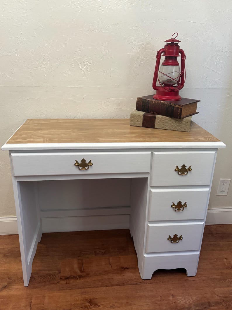 May include: White desk with four drawers and a wood top. The desk has gold-colored handles. A red lantern and books are on top of the desk.