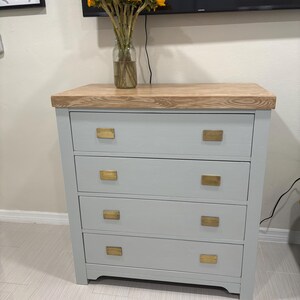 May include: A light gray dresser with four drawers and gold-colored rectangular drawer pulls. The dresser has a wood top and sits on a light-colored wood floor.