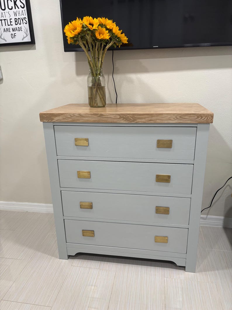 May include: A light gray dresser with four drawers and gold-colored rectangular drawer pulls. The dresser has a wood top and sits on a light-colored wood floor.