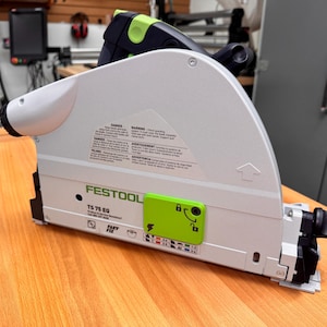 Könnte beinhalten: Eine weiße und grüne Festool TS 75 EQ-Säge mit schwarzem Griff und grünem Sicherheitsaufkleber. Die Säge liegt auf einer Holzoberfläche.