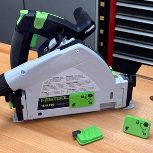 Op de afbeelding: Een groene en zwarte Festool TS 55 FEQ-zaag met een stofafzuiging en een groene veiligheidsschakelaar. De zaag staat op een houten oppervlak met twee groene veiligheidssleutels.