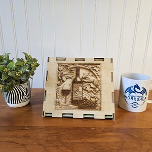 May include: A wooden box with a laser-engraved design of a wine bottle, grapes, and vines. The box is standing on a wooden table with a white mug that says "Burnt Wood Workshop" in blue lettering.