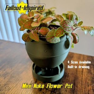 May include: A green, bomb-shaped flower pot with a plant featuring red and green leaves. The pot has a built-in drainage system and is labeled "Mini Nuke Flower Pot" and "Fallout-Inspired". Four sizes are available.