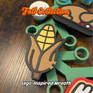 May include: A fall-themed Lego-inspired wreath with a corn on the cob design. The image features a brown, yellow, and black corn design, along with other fall-themed elements. The text "Fall Collection" and "Lego-Inspired Wreath" are also visible.