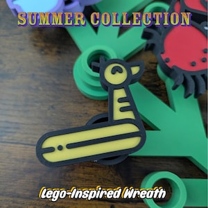 May include: A yellow and black giraffe-shaped charm with a long neck and rounded head. The text SUMMER COLLECTION appears at the top, and the text Lego-Inspired Wreath appears at the bottom. Other colorful plastic shapes are visible in the background.