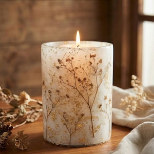 Peut inclure: Une bougie pilier blanche allumée avec un motif floral. La bougie présente des brins séchés et des accents de feuilles d'or incrustés dans la cire. La bougie est posée sur une surface en bois, avec un tissu doux de couleur neutre en arrière-plan.