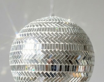 May include: A silver disco ball with a mosaic tile pattern, hanging from a thin string. The spherical ornament is covered in small, reflective tiles arranged in a chevron design, creating a dazzling effect. Ideal for parties and events.