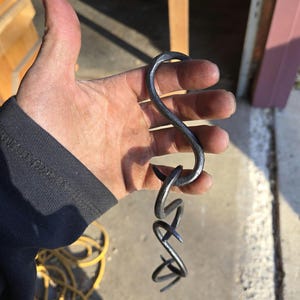 Puede incluir: Gancho de metal oscuro forjado a mano con un diseño retorcido único. El gancho presenta una parte superior curvada y una serie de bucles entrelazados en la parte inferior. El metal tiene una superficie ligeramente texturizada, lo que sugiere una calidad artesanal. El gancho se sostiene en una mano.