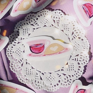 May include: A white doily with a pink teacup and saucer with a pink heart-shaped cookie on a plate. The teacup is filled with pink liquid.