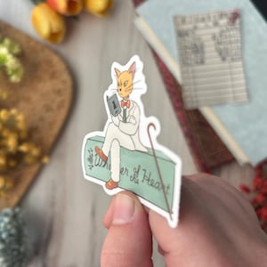 May include: A sticker featuring a cat-like character in a white suit, reading a book and holding a cane. The sticker has the text "Whisper of the Heart" on a light green surface. The cat has orange fur and a red bow tie.