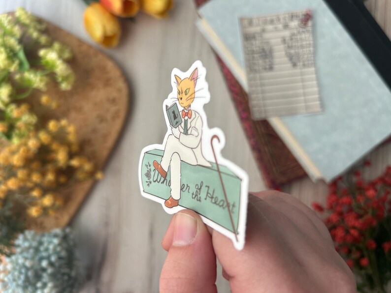 May include: A sticker featuring a cat-like character in a white suit, reading a book and holding a cane. The sticker has the text "Whisper of the Heart" on a light green surface. The cat has orange fur and a red bow tie.