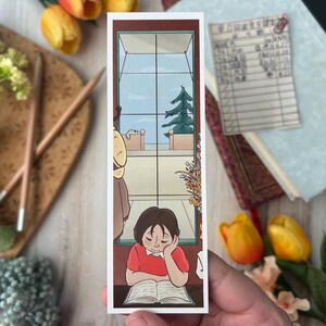 May include: A rectangular bookmark featuring an illustration of a person reading a book by a window. The person is wearing a red shirt and resting their head on their hand. The window shows a view of a tree and sky.
