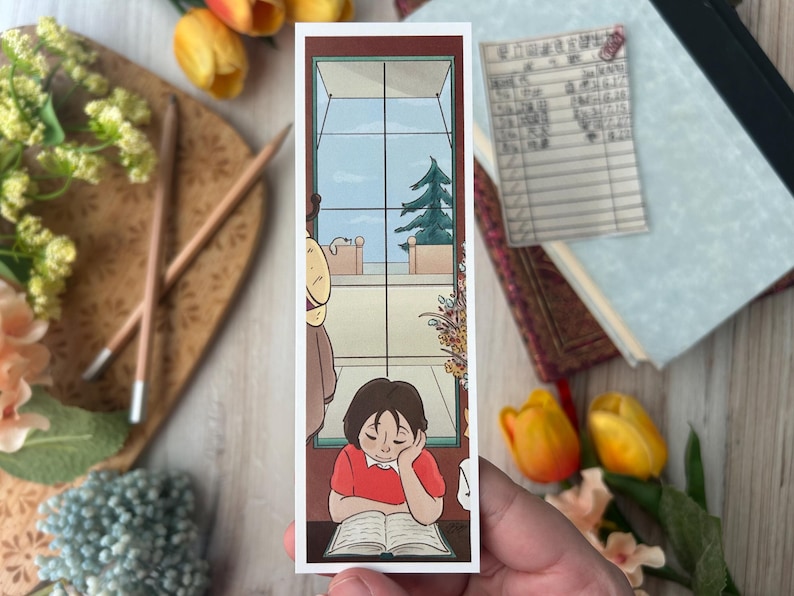 May include: A rectangular bookmark featuring an illustration of a person reading a book by a window. The person is wearing a red shirt and resting their head on their hand. The window shows a view of a tree and sky.