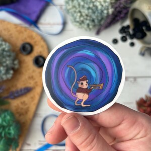 May include: A round sticker featuring a cartoon mouse playing a trumpet. The mouse is wearing a hat and jacket, set against a swirling blue and purple background. The sticker is held in a hand.