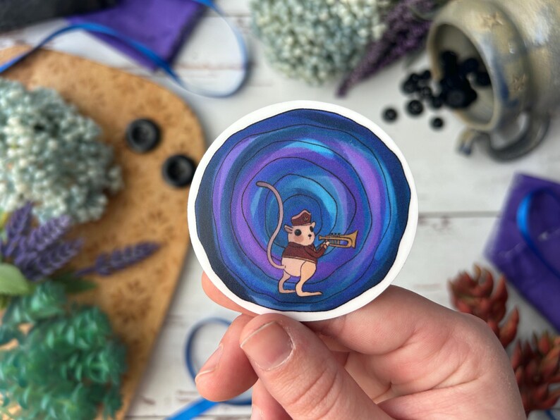 May include: A round sticker featuring a cartoon mouse playing a trumpet. The mouse is wearing a hat and jacket, set against a swirling blue and purple background. The sticker is held in a hand.