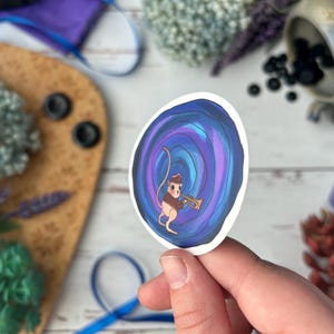 May include: A whimsical sticker featuring a cartoon mouse playing a trumpet. The mouse is dressed in a brown vest and hat, set against a swirling blue and purple background. The sticker is held in a hand, with a blurred background of decorative items.