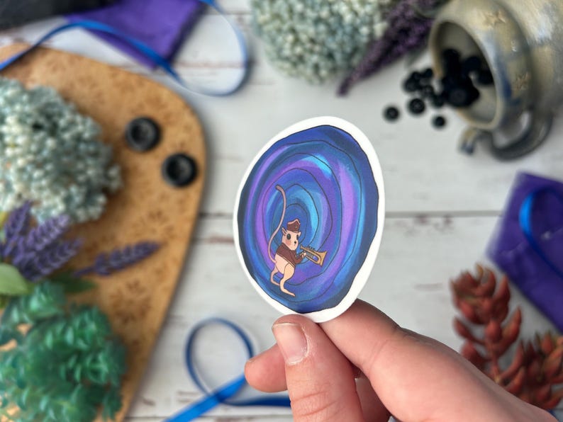 May include: A whimsical sticker featuring a cartoon mouse playing a trumpet. The mouse is dressed in a brown vest and hat, set against a swirling blue and purple background. The sticker is held in a hand, with a blurred background of decorative items.