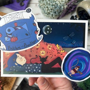 May include: A hand holds a collection of stickers and a print. The print features a girl with a black cat under a night sky with dragonflies. Stickers include a blue cartoon character, and a mouse playing a trumpet.