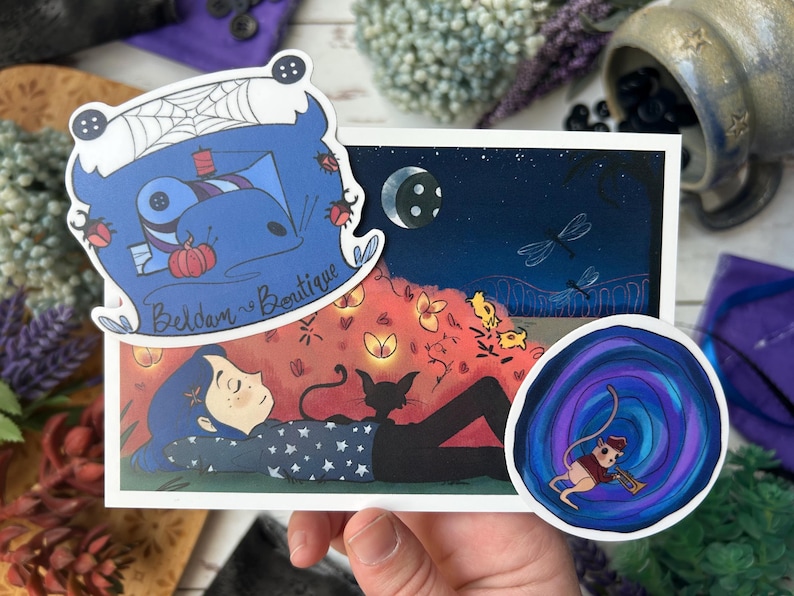 May include: A hand holds a collection of stickers and a print. The print features a girl with a black cat under a night sky with dragonflies. Stickers include a blue cartoon character, and a mouse playing a trumpet.
