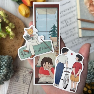 May include: A collection of stickers featuring illustrations of a cat reading a book, a person reading, and a couple with a bicycle. The cat sticker has the text "Whisper of the Heart". The stickers are in a hand.