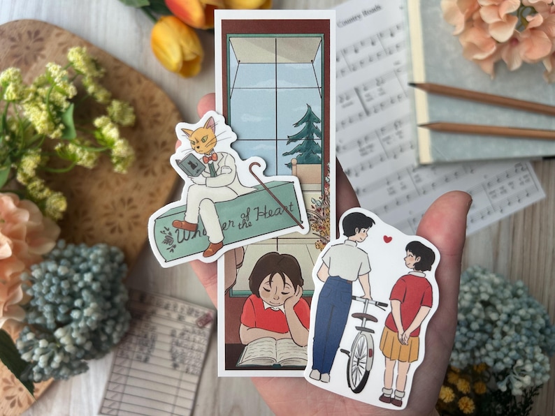 May include: A collection of stickers featuring illustrations of a cat reading a book, a person reading, and a couple with a bicycle. The cat sticker has the text "Whisper of the Heart". The stickers are in a hand.