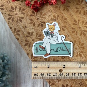 May include: A sticker featuring a cat character in a white suit, reading a book and holding a cane. The sticker has the text "Whisper of the Heart" and is approximately 3 inches long.