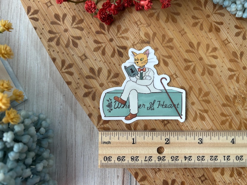 May include: A sticker featuring a cat character in a white suit, reading a book and holding a cane. The sticker has the text "Whisper of the Heart" and is approximately 3 inches long.