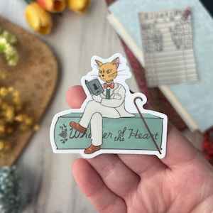 May include: A sticker featuring a cartoon cat in a white suit, reading a book. The cat is sitting on a teal-colored box with the words "Whisper of the Heart" written on it. The cat has orange fur and is wearing a bow tie.