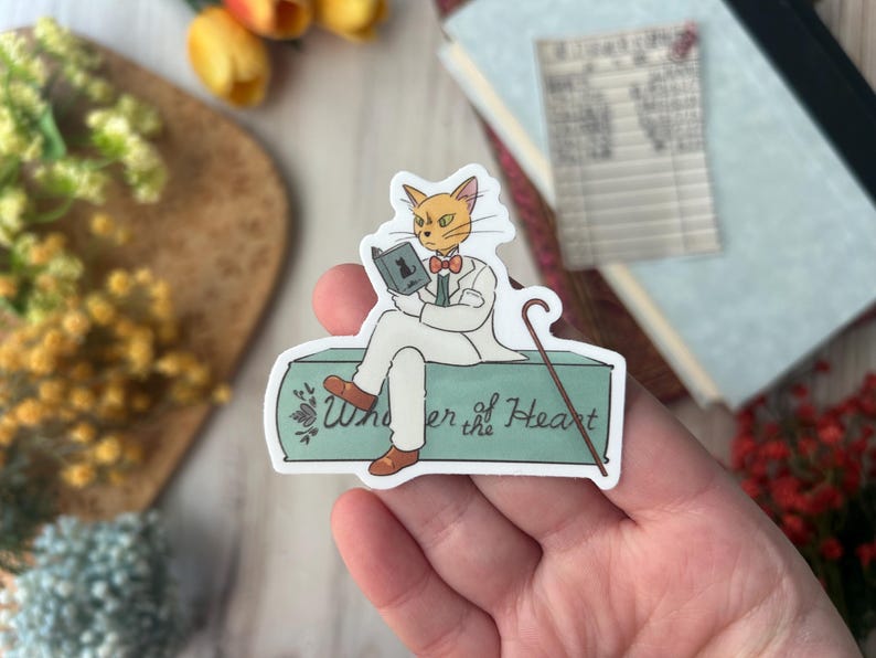 May include: A sticker featuring a cartoon cat in a white suit, reading a book. The cat is sitting on a teal-colored box with the words "Whisper of the Heart" written on it. The cat has orange fur and is wearing a bow tie.