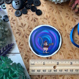 May include: A circular sticker featuring a cartoon mouse playing a trumpet, set against a swirling blue and purple background. The sticker is approximately 2 inches in diameter. The image includes a wooden ruler, buttons, and blue ribbon.