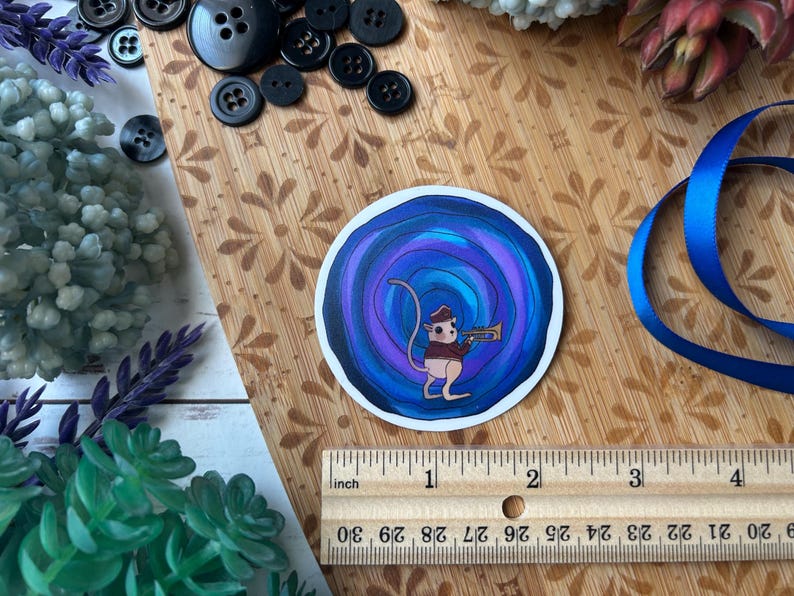May include: A circular sticker featuring a cartoon mouse playing a trumpet, set against a swirling blue and purple background. The sticker is approximately 2 inches in diameter. The image includes a wooden ruler, buttons, and blue ribbon.