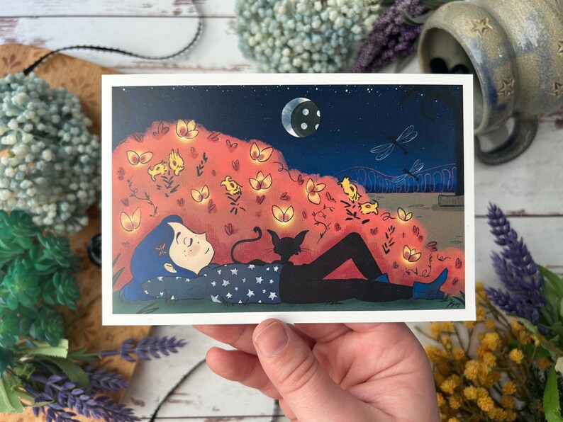 May include: A whimsical art print depicts a person with blue hair and a starry shirt, lying beneath a night sky with a crescent moon and fireflies. A black cat rests beside them. The illustration features a vibrant color palette and a dreamlike quality.