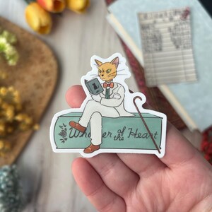 May include: A sticker featuring a cat dressed in a white suit, reading a book, and sitting on a teal surface with the text "Wonder of the Heart." The cat has orange fur, a bow tie, and a cane.