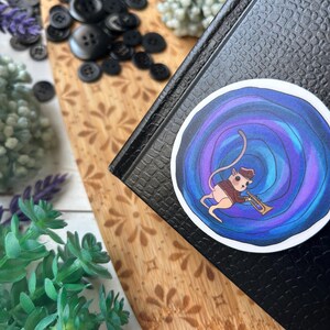 May include: A round sticker featuring a cartoon mouse playing a trumpet. The mouse is dressed in a brown vest and hat, set against a swirling blue and purple background. The sticker is placed on a black textured surface.