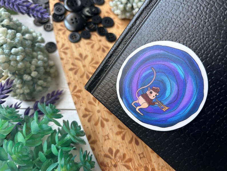 May include: A round sticker featuring a cartoon mouse playing a trumpet. The mouse is dressed in a brown vest and hat, set against a swirling blue and purple background. The sticker is placed on a black textured surface.